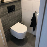 Een moderne badkamer met een wandtoilet, donkergrijze tegelwand, grijze vloertegels, een zwarte handdoek bij een kleine wastafel en de Branco RVS toiletborstel van yessential.nl in zijn stijlvolle houder naast het toilet.