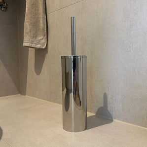 De Juano Chroom WC Set van yessential.nl, met een roestvrijstalen toiletborstelhouder met een lange steel, staat op de beige tegelvloer bij de muur. Boven coördineert een beige handdoek met andere chromen toiletaccessoires in de badkamer.