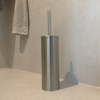 De Tapio RVS WC Set (no-drill) van yessential.nl staat op een lichtgrijs betegelde badkamervloer, met een hangende handdoek op de achtergrond, waardoor een elegante en moderne toiletaccessoires look ontstaat.