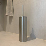 De Ludo RVS WC Set (no-drill) van yessential.nl, met een roestvrijstalen toiletborstelhouder met bijpassende handgreep, is geplaatst op een licht betegelde badkamervloer in de buurt van een grijze handdoek die aan de muur hangt.
