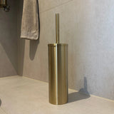 De Ludo Gold WC Set (no-drill) van yessential.nl, met een goudkleurige toiletborstelhouder met bijpassende handgreep, staat op lichtbeige tegels. Op de achtergrond hangt een grijze handdoek aan de muur.