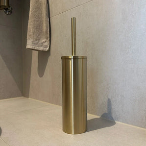 De Juano Gold WC Set (no-drill) van yessential.nl, met een gouden toiletborstel en cilindervormige houder, staat op lichte badkamertegels met een grijze handdoek op de achtergrond.