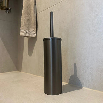 De Tygo Gunmetal WC Set (no-drill) van yessential.nl heeft een metalen toiletborstelhouder met bijpassende handgreep, die op lichte tegels bij de muur staat en wordt aangevuld met een nabijgelegen beige handdoek.
