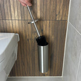 Een hand houdt de yessential.nl Extra Borstel met vervangbare kop boven een roestvrijstalen houder op een wand met houten panelen, naast een toilet met witte zitting en een betegelde vloer - klaar om schoongemaakt te worden.
