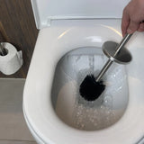 Een hand met de yessential.nl Extra Borstel, voorzien van een verwisselbare kop, reinigt de binnenkant van een witte toiletpot onder stromend water; een toiletpapierrol is te zien op een nabijgelegen houder.