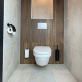Moderne minimalistische badkamer met wandgemonteerd toilet, wand met houten panelen, ingebouwde toiletpapierhouder links en de Branco Black toiletborstel zwart van yessential.nl rechts. De lichtgrijze tegelvloer en -wanden maken de look compleet.