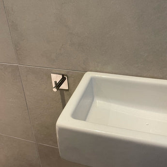 Een witte rechthoekige wastafel is gemonteerd op een betegelde muur met de Ludo Chroom WC Set (no-drill) van yessential.nl, inclusief een metalen toiletpapierhouder die ongewoon dicht bij de rand van de wastafel is geïnstalleerd voor eenvoudige installatie zonder boren.