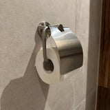 Een wc-papierrol is aan de muur bevestigd onder de glanzende Julio RVS houder van yessential.nl, een slanke metalen houder die is bevestigd aan lichtgekleurde betegelde badkamermuren.