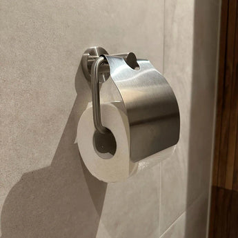 Een wc-papierrol is aan de muur bevestigd onder de glanzende Julio RVS houder van yessential.nl, een slanke metalen houder die is bevestigd aan lichtgekleurde betegelde badkamermuren.