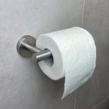 Een rol toiletpapier hangt aan een Tino RVS toiletrolhouder van yessential.nl, gemonteerd op een lichtgrijze betegelde muur. Het reliëfpapierpatroon en de moderne houder voegen stijl toe naast andere strakke toiletaccessoires.