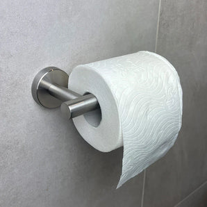 Een rol toiletpapier hangt aan een Tino RVS toiletrolhouder van yessential.nl, gemonteerd op een lichtgrijze betegelde muur. Het reliëfpapierpatroon en de moderne houder voegen stijl toe naast andere strakke toiletaccessoires.