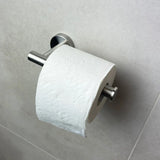 Een rol toiletpapier met een golvend en gebloemd reliëfpatroon zit op de Tino RVS van yessential.nl, gemonteerd op een lichtgrijze betegelde muur. Deze stijlvolle houder geeft je collectie toiletaccessoires een elegante uitstraling.
