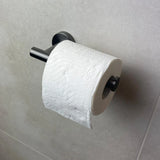 Een rol toiletpapier met een golvend reliëfpatroon hangt aan de Tino Gunmetal toiletrolhouder van yessential.nl, gemonteerd op een lichtgrijze betegelde muur voor een strakke, moderne toevoeging aan je badkameraccessoires.