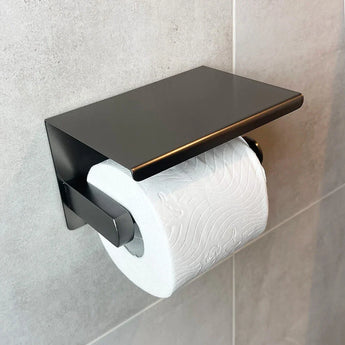 De Tygo Gunmetal WC Set (no-drill) van yessential.nl is een moderne gunmetal toiletpapierhouder met een plat bovenblad, gemonteerd zonder boren op een lichtgrijs betegelde badkamermuur en met een rol wit toiletpapier.