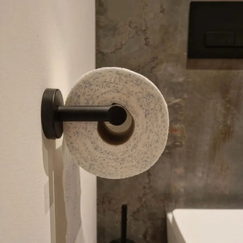 De Romano Black WC-set van yessential.nl heeft een wc-papierrol met donkere vlekken op een matzwarte houder, gemonteerd tegen een witte muur en een rustiekgrijze getextureerde achtergrond.
