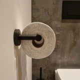 Het Tino Black toiletpapier van yessential.nl hangt aan een RVS houder tegen een witte muur, gecombineerd met matzwarte accessoires in een moderne badkamer met een betonnen achtergrond.