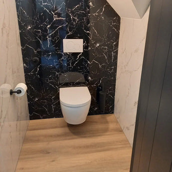 Een moderne badkamer met het yessential.nl Tino Black wandcloset, zwart marmeren achterwand, witte muren, houten vloer, matzwarte accessoires en een RVS toiletrolhouder aan de linkermuur.