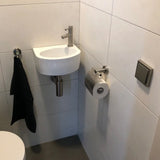 Een kleine witte wastafel met een zilveren kraan staat in een betegelde hoek boven een grijze vloer. Een zwarte handdoek hangt in de buurt en een yessential.nl Julio RVS toiletrolhouder is vlak bij de wastafel gemonteerd. Aan de muur hangt een vierkante spoelknop.