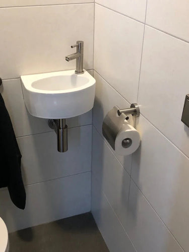 Een kleine witte wastafel met een zilveren kraan staat in een betegelde hoek boven een grijze vloer. Een zwarte handdoek hangt in de buurt en een yessential.nl Julio RVS toiletrolhouder is vlak bij de wastafel gemonteerd. Aan de muur hangt een vierkante spoelknop.