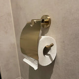 Een Mano Gold WC-set toiletpapierhouder van yessential.nl is gemonteerd op een beige muur, met de rol verkeerd geïnstalleerd zodat het papier van de muur af hangt in plaats van onder het deksel.
