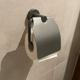 De Julio Pearl Grey wc-rolhouder van yessential.nl, een moderne toiletaccessoire in parelgrijs, is aan de beige badkamermuur bevestigd en bedekt het papier deels met een stijlvolle metalen klep.