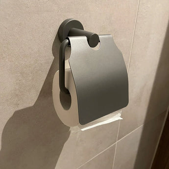 De Julio Pearl Grey wc-rolhouder van yessential.nl, een moderne toiletaccessoire in parelgrijs, is aan de beige badkamermuur bevestigd en bedekt het papier deels met een stijlvolle metalen klep.