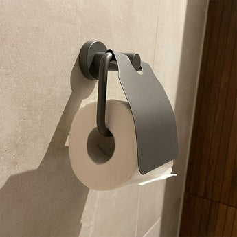 Een rol toiletpapier zit op de yessential.nl Julio Pearl Grey toiletrolhouder met klep, gemonteerd op een betegelde muur. Rechts is een houten paneel zichtbaar.