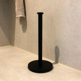 De Delo Black WC Set (no-drill) van yessential.nl, een strakke matzwarte papieren handdoekhouder, staat stijlvol op een lichte badkamervloer bij de muur, met een beige handdoek op de achtergrond.