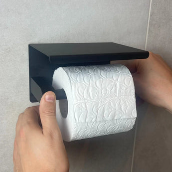 De handen van een persoon plaatsen een rol wit toiletpapier met reliëf op de Juano Black WC Set (no-drill) van yessential.nl, die zonder boren wordt gemonteerd op een lichtgekleurde betegelde muur.