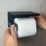 De handen van een persoon plaatsen een rol toiletpapier op een yessential.nl Tazio Black (No Drill) toiletrolhouder die gemonteerd is op een betegelde badkamermuur.