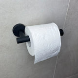 Een rol toiletpapier met een golf- en bloemmotief in reliëf hangt aan een Tino Black toiletrolhouder van yessential.nl, bevestigd aan een lichtgrijze betegelde muur.