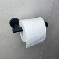 Een rol toiletpapier met een golf- en bloemmotief in reliëf hangt aan een Tino Black toiletrolhouder van yessential.nl, bevestigd aan een lichtgrijze betegelde muur.