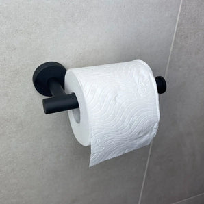 Een rol toiletpapier met een golf- en bloemmotief in reliëf hangt aan een Tino Black toiletrolhouder van yessential.nl, bevestigd aan een lichtgrijze betegelde muur.