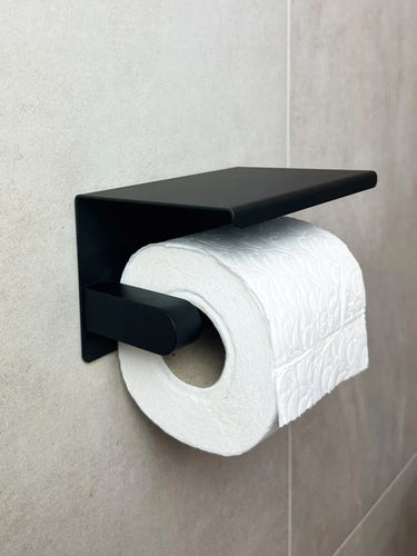 Een rol wit toiletpapier met reliëf op een yessential.nl Tazio Black toiletrolhouder met planchet, bevestigd aan een betegelde badkamerwand.