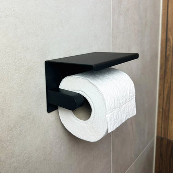 Een rol wit toiletpapier met reliëf op een yessential.nl Tazio Black toiletrolhouder met planchet, bevestigd aan een betegelde badkamerwand.