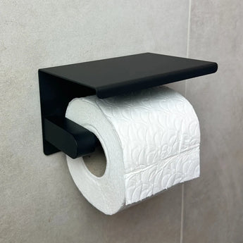 Een rol wit toiletpapier met reliëf is gemonteerd op de Tazio Black toiletrolhouder van yessential.nl, die is bevestigd aan een lichtgekleurde betegelde muur.