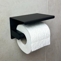 Een rol wit toiletpapier met reliëf is gemonteerd op de Tazio Black toiletrolhouder van yessential.nl, die is bevestigd aan een lichtgekleurde betegelde muur.