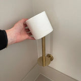 Een hand plaatst een rol toiletpapier op de Roberto Gold toiletrolhouder van yessential.nl, een schuine, goudkleurige houder die aan de badkamermuur is bevestigd - ideaal voor het opwaarderen van je badkameraccessoires.