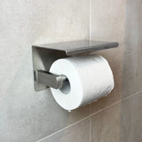 Een rol toiletpapier op de yessential.nl Ludo RVS WC Set (no-drill), een moderne wandhouder met planchet, is bevestigd aan een lichtgrijs betegelde badkamermuur.