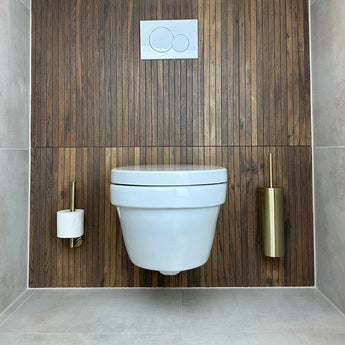 Een moderne badkamer met een wit wandcloset, verticale houten lambrisering en de Cosimo Gold WC-set van yessential.nl: gouden toiletborstel rechts en toiletpapierhouder links. De doorspoelknop zit boven het toilet.