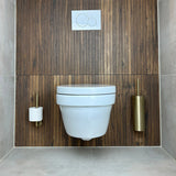 Een moderne badkamer met een aan de muur bevestigd wit toilet, houten lambrisering en de yessential.nl Romano Gold WC-set - met rechts een gouden borstelhouder en links een papierhouder - afgewerkt met lichtgrijze tegels.