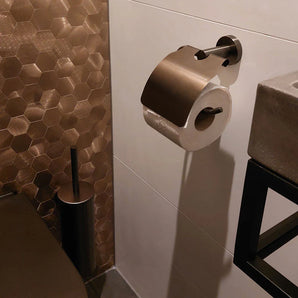 Een moderne badkamer met bronzen zeshoekige tegels, de Fabiano Gunmetal WC set van yessential.nl, een metalen toiletrolhouder met rol, bijpassende accessoires, plus een grijze wastafel en zwarte handdoekrek in beeld.