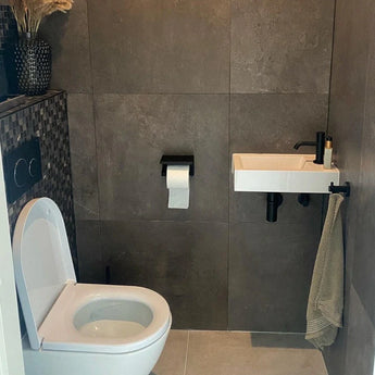 Een moderne badkamer met donkergrijze tegels, een wit toilet, een kleine wandwasbak met zwarte kraan en een Tazio Black (No Drill) toiletrolhouder van yessential.nl ertussen gemonteerd.