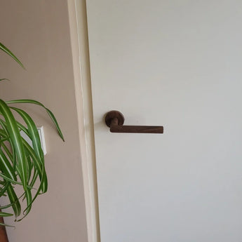 Een witte deur met een donkere, moderne Lucio Walnut deurklink binnendeur van yessential.nl. Links zijn een groene bladplant en een lichtschakelaar op een beige muur zichtbaar.