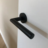 De Lucio Zwarte deurklink van yessential.nl is gemonteerd op een witte deur, met een beige muur en houten vloer op de achtergrond. Dit strakke zwarte deurkrukken ontwerp brengt moderne stijl in elk interieur.