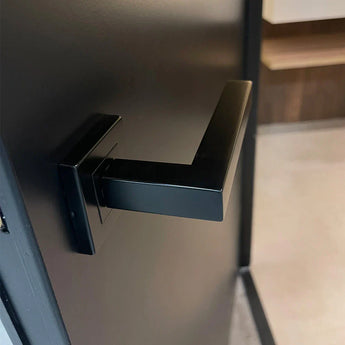 Een close-up van de moderne Domaso Black deurklink van yessential.nl, met een vierkante basis en minimalistisch hoekig ontwerp, gemonteerd op een donkere deur met daarachter de zacht vervaagde kamer zichtbaar.