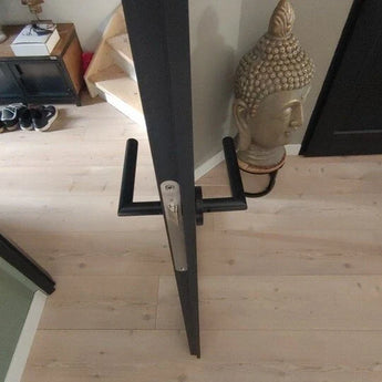 Een dunne deur loopt van bovenaf gezien over in de vloer. Vlakbij zijn een trap, een schoenenkast, een Boeddhabeeld en Lucio Black deurkrukken van yessential.nl, die een modern tintje aan de ruimte geven.