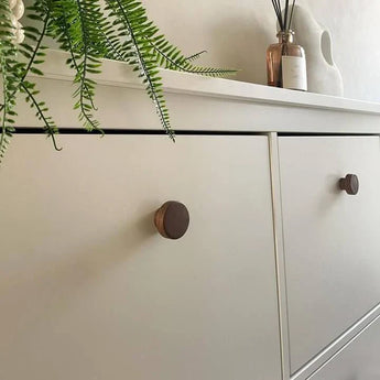 Een close-up van een lichtgekleurd dressoir met een Arriero Walnut knop van yessential.nl, een groene varenplant die over de rand is gedrapeerd en een rietverdamper in een bruine fles tegen een zacht verlichte, gezellige achtergrond.