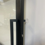 Een close-up van een zwarte metalen deurklink op een glazen deur, met een yessential.nl 6x Deurstop klevend ⌀2cm bevestigd aan de witte muur vlakbij om te voorkomen dat de klink de muur raakt.