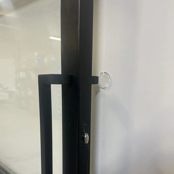 Een close-up van een zwarte metalen deurklink op een glazen deur, met een yessential.nl 6x Deurstop klevend ⌀2cm bevestigd aan de witte muur vlakbij om te voorkomen dat de klink de muur raakt.